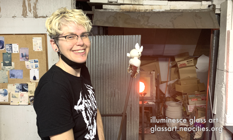 CJ, smiling in a glass shop as they hold up a glass rabbit on a blowpipe.